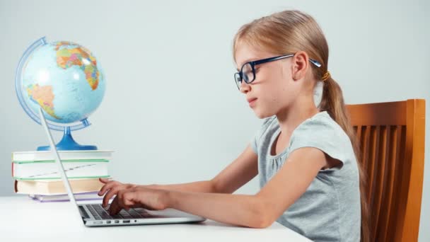 Estudiante de retrato de 7-8 años escribiendo texto en el portátil y sentado en la mesa y sonriendo a la cámara con los dientes. Aislado sobre blanco — Vídeos de Stock