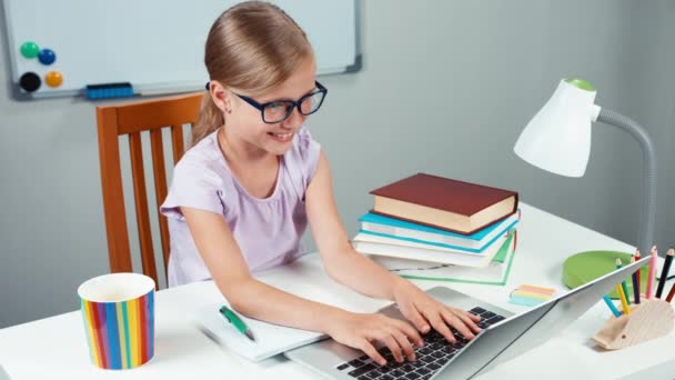 Retrato estudiante niña 7-8 años haciendo la tarea con el ordenador portátil y la escritura en su cuaderno de ejercicios. Pulgar hacia arriba. Ok. — Vídeos de Stock