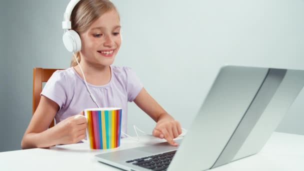 Πορτρέτο φοιτητής κορίτσι παιδί 7-8 χρόνια χρήση φορητού υπολογιστή, ακούγοντας μουσική στα ακουστικά και κρατώντας το φλιτζάνι του τσαγιού — Αρχείο Βίντεο
