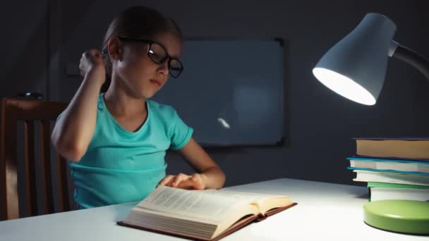 Σχολείο κορίτσι 7-8 χρόνια χασμουρητά και διαβάζοντας ένα βιβλίο τη νύχτα στο γραφείο της — Αρχείο Βίντεο