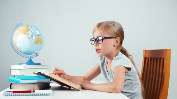 Szkoła dziewczyna cztery oczy czytając jej podręcznika i uśmiecha się do kamery. Dziecko siedzi przy biurku na białym tle — Wideo stockowe