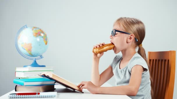 Chica de la escuela de cuatro ojos leyendo libro de texto y comer croissant. Niño sentado en el escritorio aislado sobre fondo blanco — Vídeos de Stock