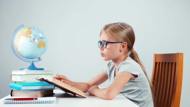 Škola dívka čtyři oči čtení učebnice a směje se na kameru. Dítě sedí u stolu izolované na bílém — Stock video