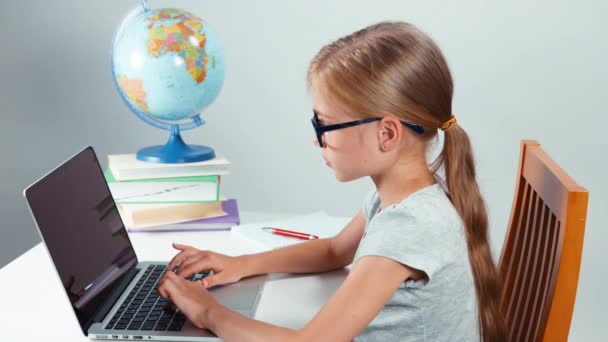学校女孩四眼打字在她的笔记本电脑和相机微笑。坐在办公桌前隔离在白色背景上的孩子。大拇指。还行 — 图库视频影像