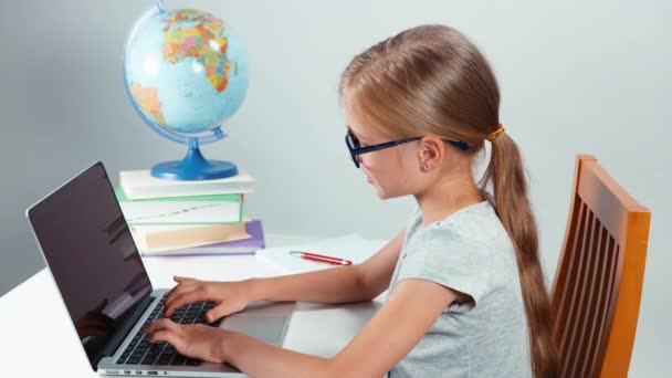 学校女孩四眼打字在她的笔记本电脑和相机微笑。孩子坐在办公桌上白色孤立。大拇指。还行 — 图库视频影像