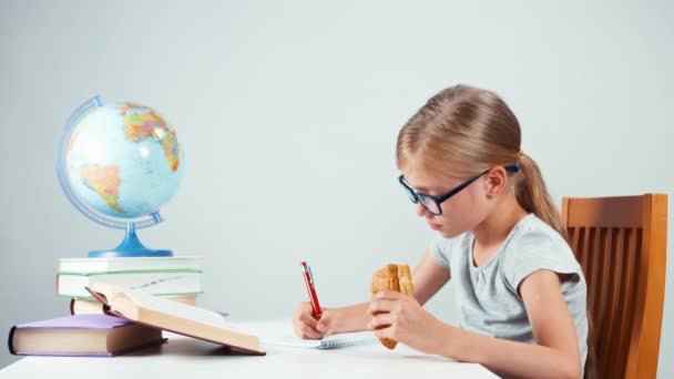 Ragazza della scuola quattro occhi che scrive nel suo quaderno e mangia croissant. Bambino seduto alla scrivania isolato su sfondo bianco — Video Stock