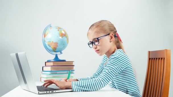 Chica de la escuela escribiendo en su computadora portátil y escribiendo en el cuaderno. Niño sentado en el escritorio aislado en blanco — Vídeos de Stock