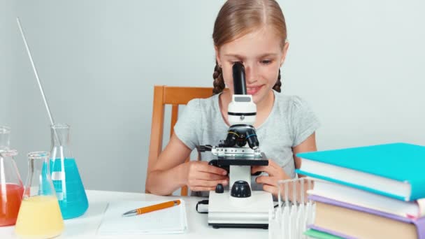 School girl using microscope and something writing in her exercise book and smiling at camera. Isolated on white — Stock Video