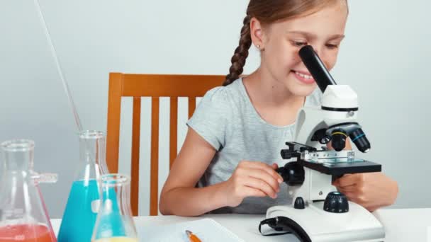 Школьница пользуется микроскопом и что-то пишет в тетрадке. Ребенок сидит за столом — стоковое видео