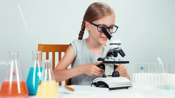 Colegiala sentada en el escritorio cerca del microscopio y sonriendo a la cámara con los dientes — Vídeos de Stock