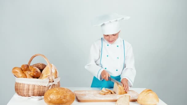 Genç baker kız 7-8 yıl ekmek kesme ve beyaz arka plan üzerinde izole mutfak masasının üzerinde bir parça yemek. Yukarı başparmak. Tamam — Stok video