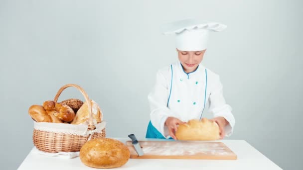 Девочка-пекарь режет буханку хлеба на кухонном столе, смотрит на камеру, изолированную на белом — стоковое видео