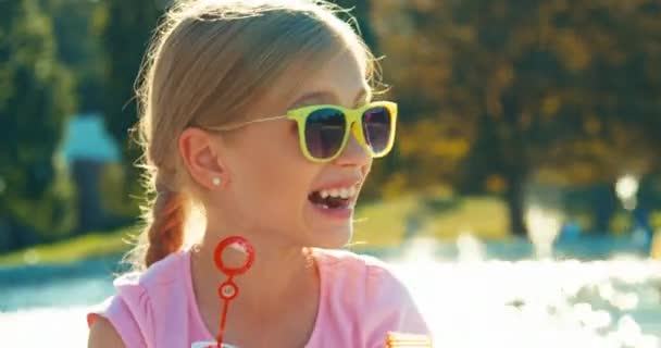 Close up portrait laughing child girl 7-8 years in sunglasses blowing soap bubbles on fountain background at sunny day — Stock Video