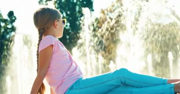 Child girl 7-8 years sitting relaxing near fountain smiling at camera and waving hand — Stock Video