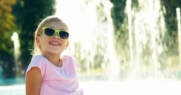 Close-up van portret meisje kind 7-8 jaar ontspannen in de buurt van fontein aan de zonnige dag en zwaaien hand op camera — Stockvideo