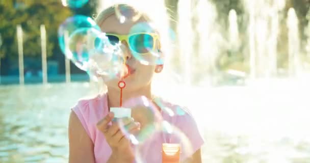 Nahaufnahme Porträt lachende Mädchen pustet Seifenblasen auf Brunnenhintergrund bei sonnigem Tag — Stockvideo
