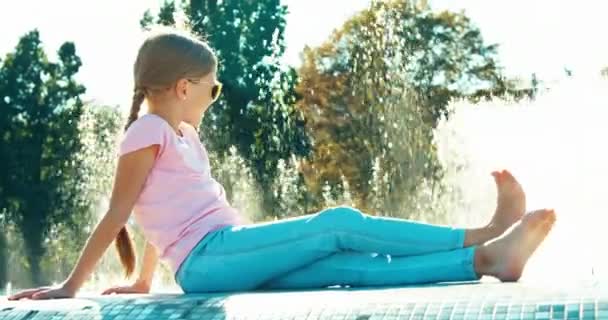 Chica 7-8 años sentado cerca de la fuente en el día soleado. Niño en gafas de sol sonriendo a la cámara — Vídeos de Stock