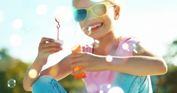 Портретна дівчина 7-8 років в сонцезахисних окулярах дме мильні бульбашки, сидячи біля фонтану і посміхаючись на камеру і махаючи рукою — стокове відео