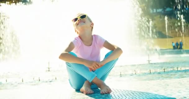 Ребенок сидит возле фонтана в солнечный день и растягивается — стоковое видео
