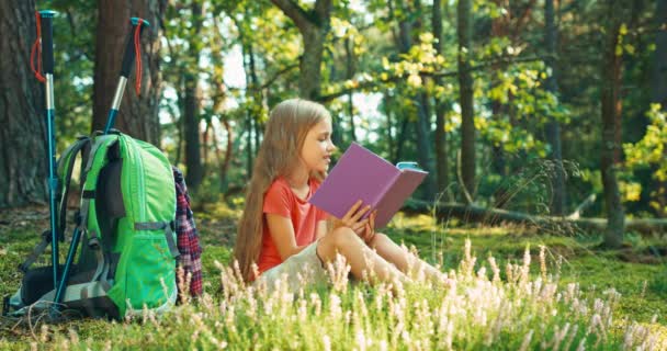 Πεζοπόρος κορίτσι παιδί 8-9 χρόνια διαβάζοντας το βιβλίο συνεδρίαση για το γρασίδι και να χαμογελά στη φωτογραφική μηχανή στο δάσος — Αρχείο Βίντεο
