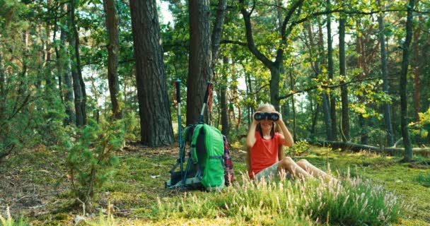 Hiker girl niño de 8-9 años usando prismáticos y mirando a la cámara en el bosque. Mano de niño saludando — Vídeos de Stock