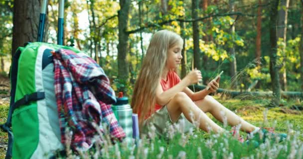 Hiker flicka barn 8-9 år med smartphone och skrattar sitter i gräset i skogen — Stockvideo