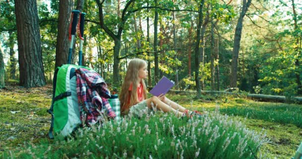 Randonneuse fille enfant 8-9 ans lire le livre et le fermer assis dans l'herbe dans la forêt et sourire à la caméra — Video