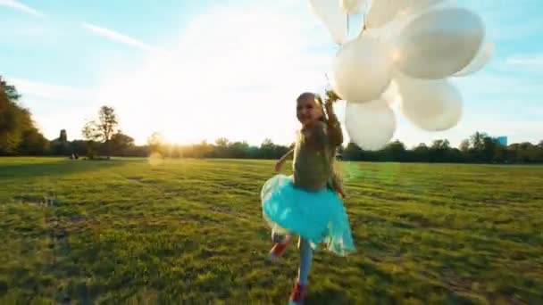 Bambino 8-9 anni che corre attraverso il campo al sole al tramonto e tiene i palloncini bianchi e sorride alla macchina fotografica — Video Stock
