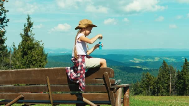 Pequena menina caminhante criança 7-8 anos soprando bolhas de sabão e sentado no banco no fundo das montanhas — Vídeo de Stock