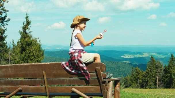 Kleines Wandermädchen 7-8 Jahre alt, spielt mit Seifenblasen und sitzt auf der Bank im Hintergrund der Berge — Stockvideo