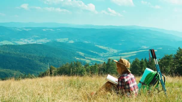 Портрет маленької дівчинки-поводиря 7-8 років читає книгу і сидить на фоні гір. Турист сів відпочити на траву — стокове відео