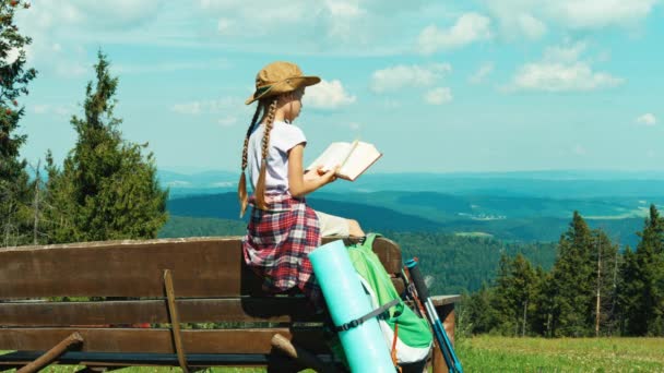 Niña excursionista niño 7-8 años leyendo libro y sentado en el banco en el fondo de las montañas — Vídeo de stock