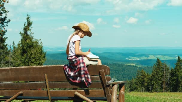 Cute little hiker girl child 7-8 years old something drawing in notebook and sitting on the bench on mountains background — Stock Video