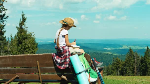 Pequeña niña excursionista viejo algo de dibujo en el cuaderno y sentado en el banco en el fondo de las montañas — Vídeos de Stock