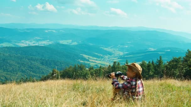 Retrato niña pequeña excursionista 7-8 años de edad mirando a la distancia a través de prismáticos y sentado en la hierba en el fondo de las montañas — Vídeo de stock