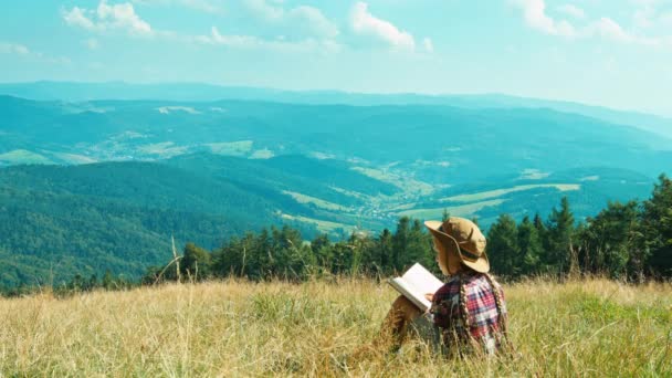 Portrét turistické dívek 7-8 let proti hory čtení knihy sedět na trávě a usmívá se na kameru Stock Video