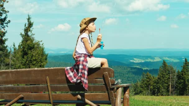 Kleines Wandermädchen 7-8 Jahre alt pustet Seifenblasen und sitzt auf der Bank im Hintergrund der Berge — Stockvideo