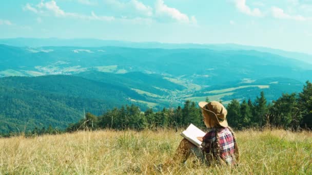 Portrait little hiker child girl 7-8 years reading book and sitting on grass on mountains background — Stock Video