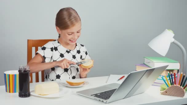 Портретная школьница 7-8 лет с помощью ножа делает сэндвич с маслом и смотрит на свой ноутбук, сидя за столом вечером — стоковое видео