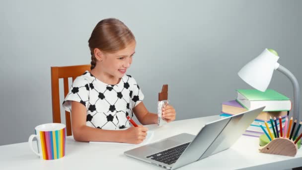 शाळेतली मुलगी तिच्या नोटबुकमध्ये लिहिते आणि संध्याकाळी डेस्कवर बसून चॉकलेट खाते आणि कॅमेरा पाहते — स्टॉक व्हिडिओ