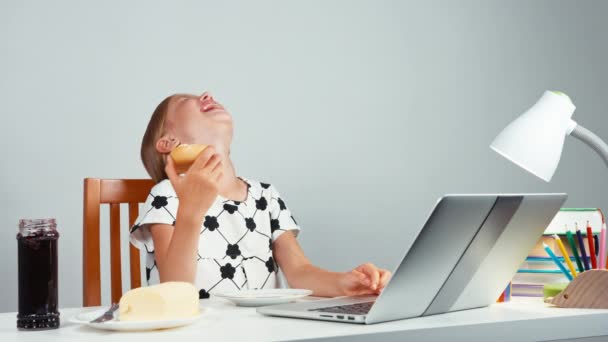 Tjej 7-8 år skrattar äter bröd med marmelad smör och använder bärbar dator sitter vid bordet isolerad på vit — Stockvideo
