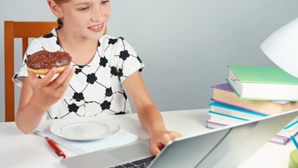 Schulmädchen isst Donut und benutzt ihren Laptop und sitzt abends am Schreibtisch — Stockvideo