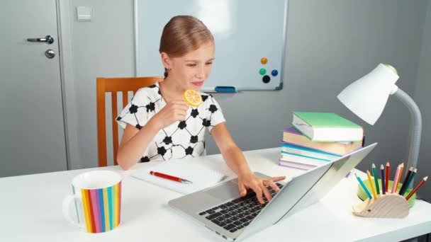 Schoolgirl menggunakan laptop dan kejutannya. Anak menjilati permen loli dan tersenyum di kamera duduk di meja di malam hari. Jempol ke atas. Baiklah. — Stok Video