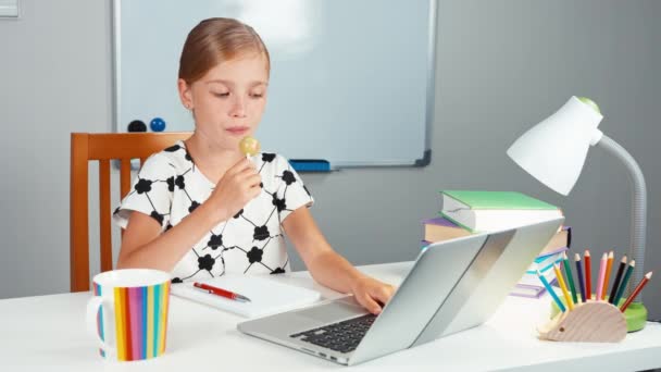 Schoolgirl using her laptop and surprise. Child licking lollipop and smiling at camera sitting at the desk in the evening — Stock Video