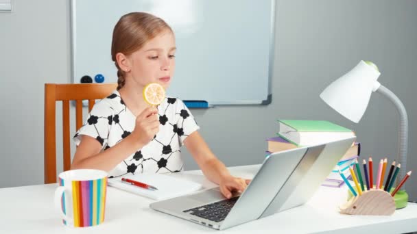 Školačka pomocí její laptop a lízání lízátko a usmívá se na kameru. Dítě sedí u stolu v večer — Stock video