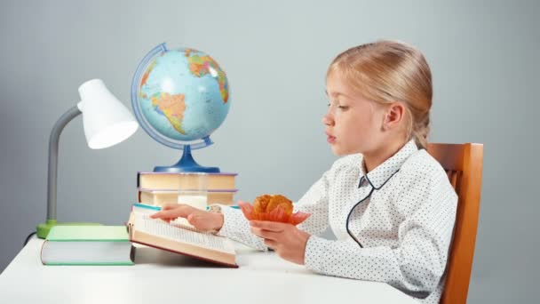 Niña de la escuela 7-8 años leyendo libro comiendo panecillo sentado en la mesa — Vídeos de Stock