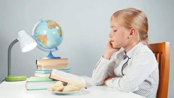 Schulmädchen isst Kartoffelchips und liest abends am Tisch Lehrbücher und blickt mit großen Augen in die Kamera — Stockvideo