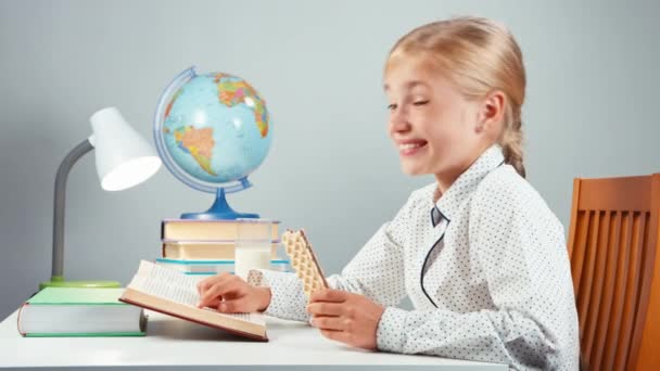 Закрыть портретную школу девочка 7-8 лет читает книгу и ест вафли и смеется сидя за столом на белом фоне — стоковое видео