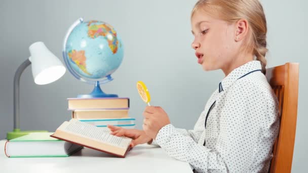 Colegiala lamiendo piruleta y asiente con la cabeza a la cámara. Niño sentado en el escritorio — Vídeos de Stock