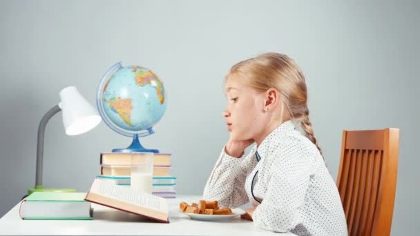 Perfil del retrato chica rubia de la escuela 7-8 años leyendo libro comiendo dulces y sentado en la mesa aislado en blanco en la noche — Vídeos de Stock